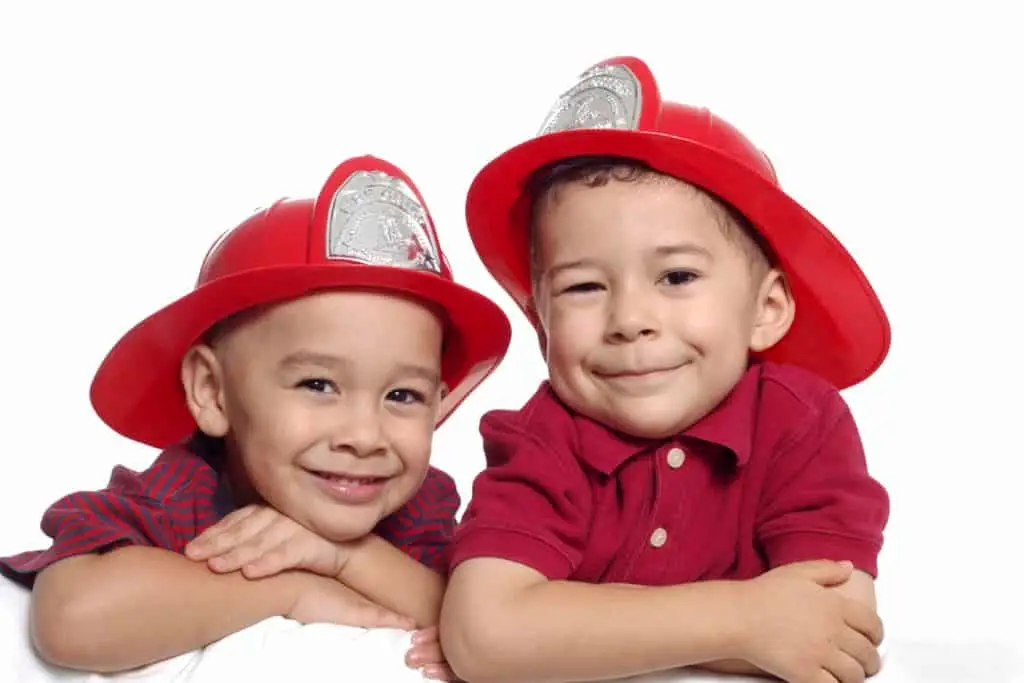 Two young children wearing red firefighter hats and smiling against a white background.