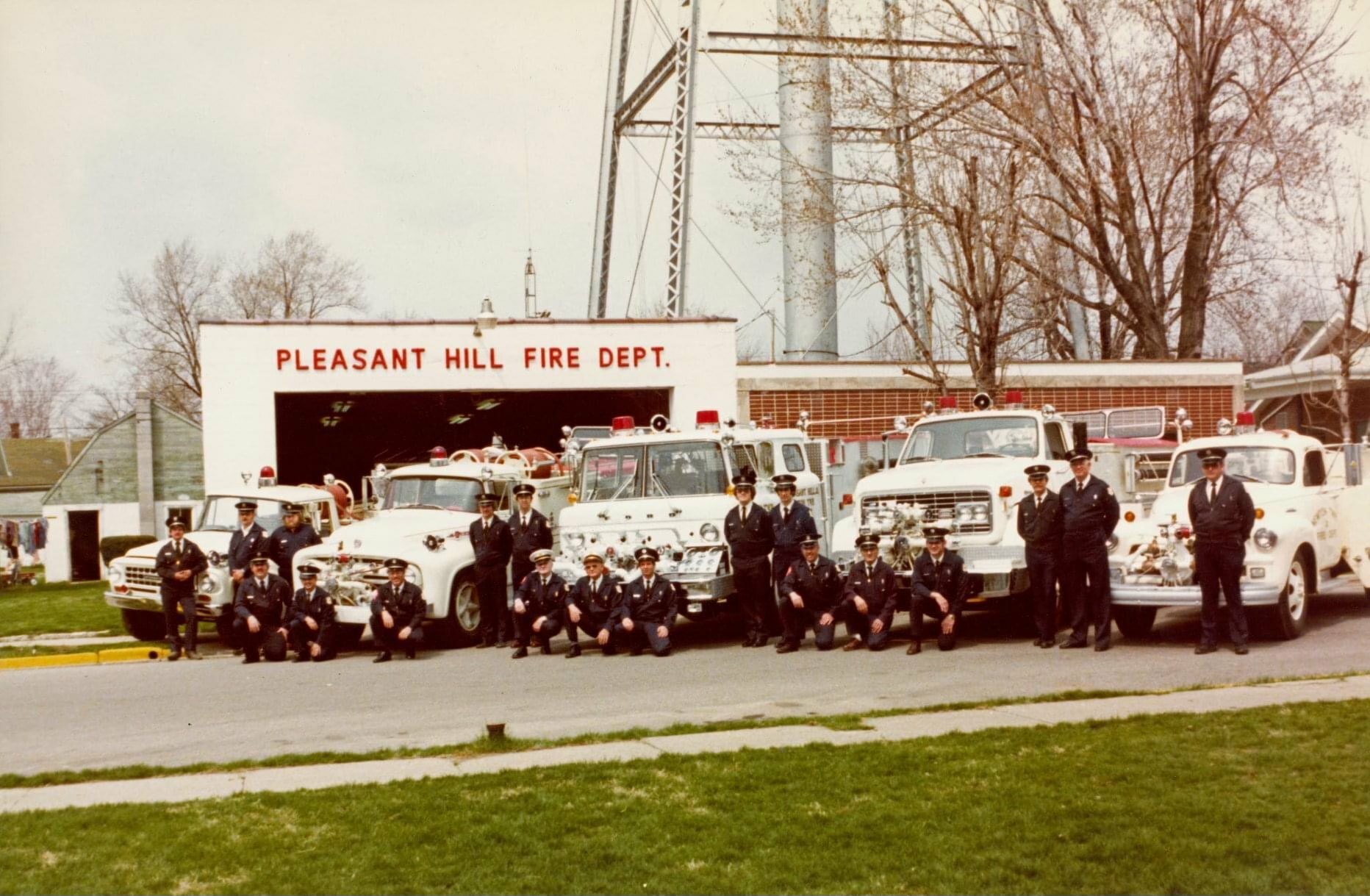 A group of firefighters in uniform pose in front of several fire trucks parked outside a building with a sign that reads "Pleasant Hill Fire Dept." Trees and a tall structure are visible in the background.