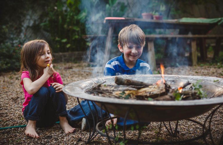 Two children sitting by a metal fire pit outdoors. The girl in a pink shirt holds a marshmallow, while the boy in a blue striped shirt watches the small fire. They are surrounded by gravel and greenery, with a table and chairs in the background.