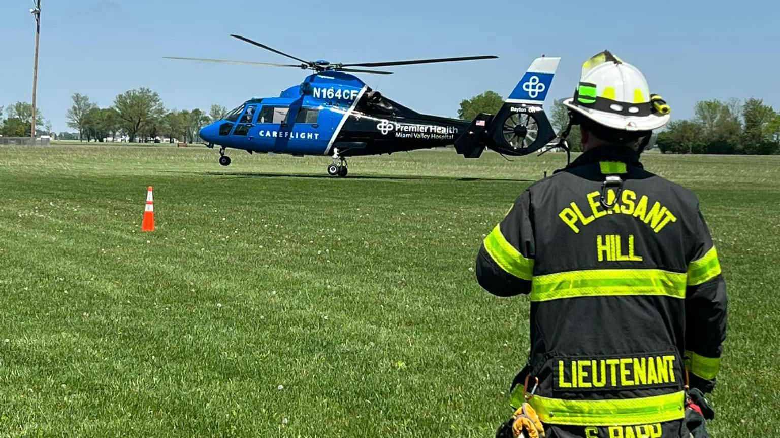 A helicopter is parked on a grassy field, displaying "Careflight" and "Premier Health" logos. In the foreground, a firefighter in a "Pleasant Hill Lieutenant" jacket stands, viewing the scene. There's a safety cone nearby.