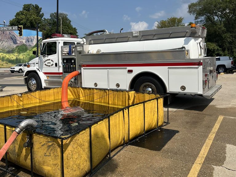 A fire truck is parked near a yellow portable water tank, with hoses connected for water transfer. The scene is set on a sunny day in a parking lot, with trees and a billboard visible in the background.