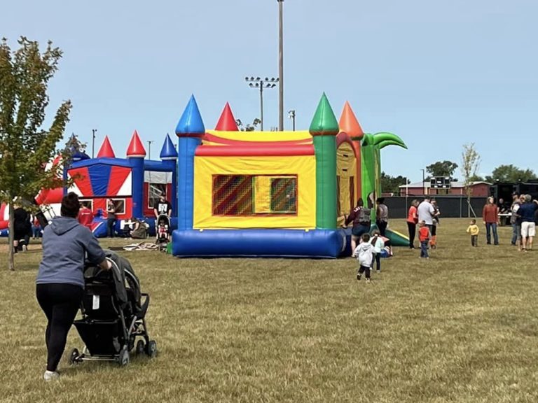 A grassy field with families enjoying a fair. A woman pushes a stroller towards colorful inflatable bounce houses. Children play nearby, and several trucks are parked in the background under a clear blue sky.