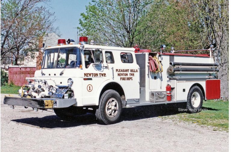 A vintage fire truck labeled "Pleasant Hill Newton Type 2 Fire Dept." is parked outdoors on a gravel surface. The truck is white with red and chrome accents and has hoses and equipment attached. Trees and a building are visible in the background.