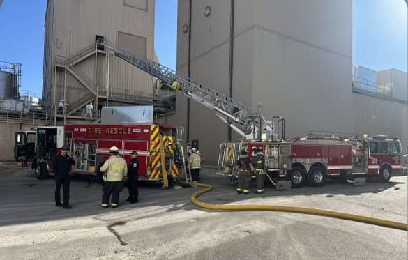 Firefighters and two fire trucks are at the scene of an emergency near a large industrial building. A ladder extends from one truck to the building. Firefighters are gathered, appearing to assess the situation.