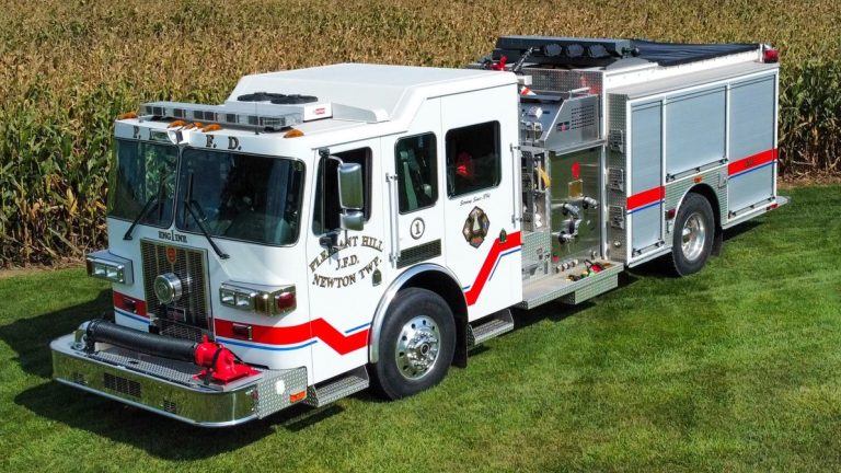 A white fire truck with red and blue stripes is parked on a grassy area. It has various equipment compartments and visible logos on its side. Cornfields are in the background.