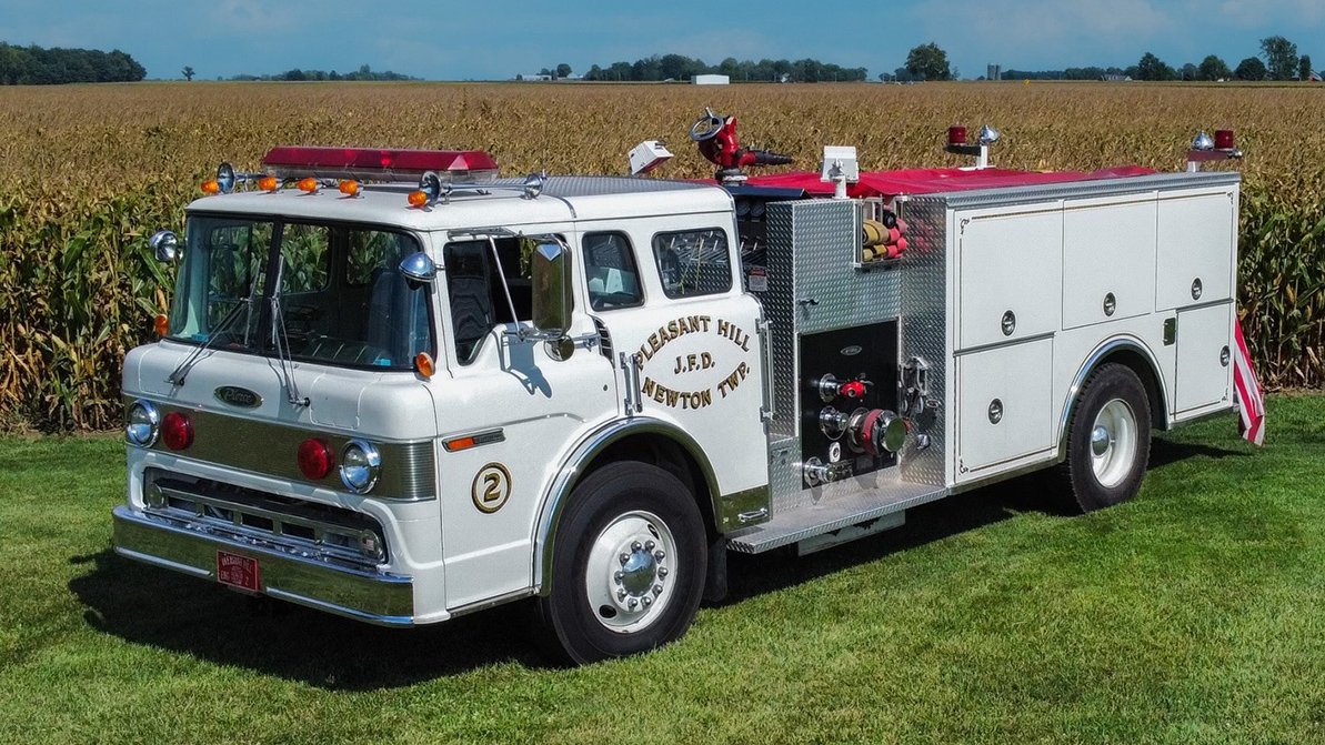 A vintage white fire truck with "Pleasant Hill Newton Twp" written on the side is parked on grassy terrain. It features red lights, a silver grille, and multiple compartments. A cornfield and blue sky are in the background.