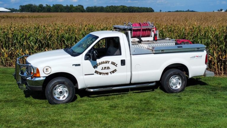 A white pickup truck labeled "Pleasant Hill V.F.D. BRUSH TRUCK" is parked on grass in front of a cornfield. The truck has firefighting equipment, including hoses and a water tank, secured in the bed. The sky is blue with a few clouds.