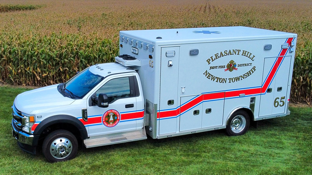 A white and silver ambulance with red and blue stripes parked on grass beside a cornfield. The vehicle is marked "Pleasant Hill Newton Township Joint Fire District" and has a medical emblem on the side.