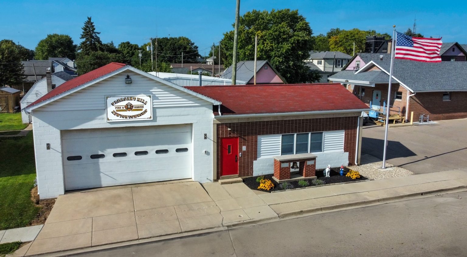 A small-town fire station with a red-roofed building. An American flag stands near the entrance. A sign above the garage door reads "Fire Dept." The street and surrounding houses are visible in the background.