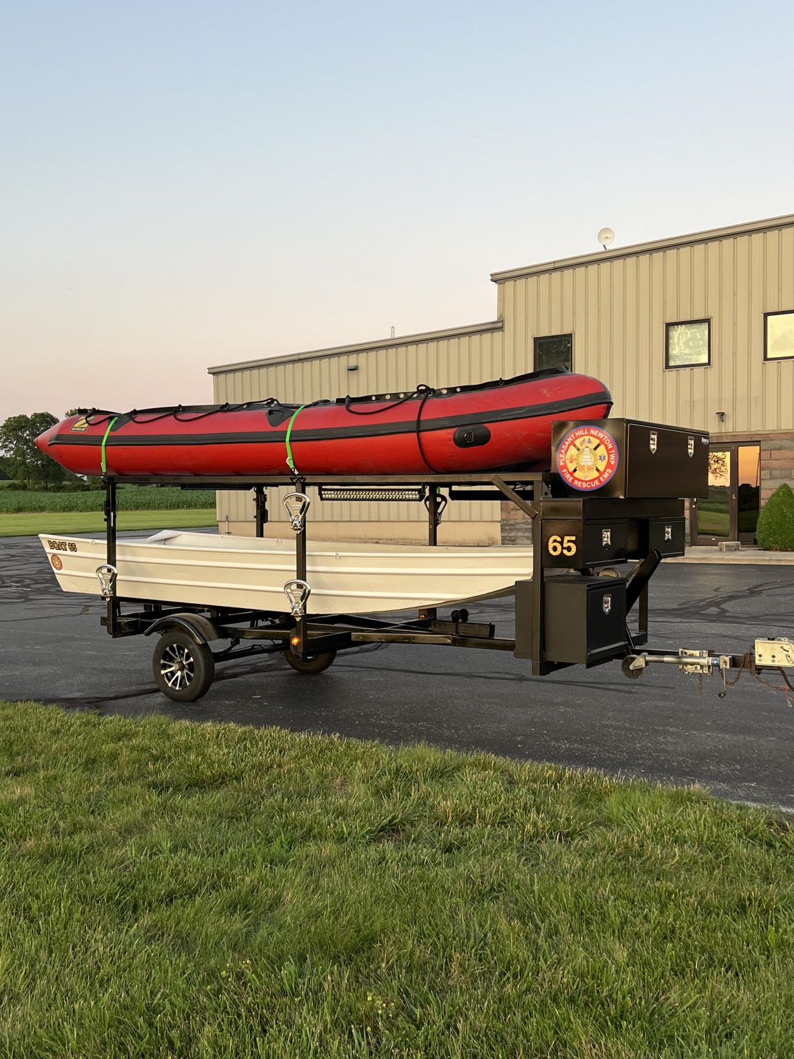 A trailer in a parking lot carries a red inflatable boat on top and a white rowboat below. The trailer is black, labeled "65," and is parked near a beige industrial building at sunset.