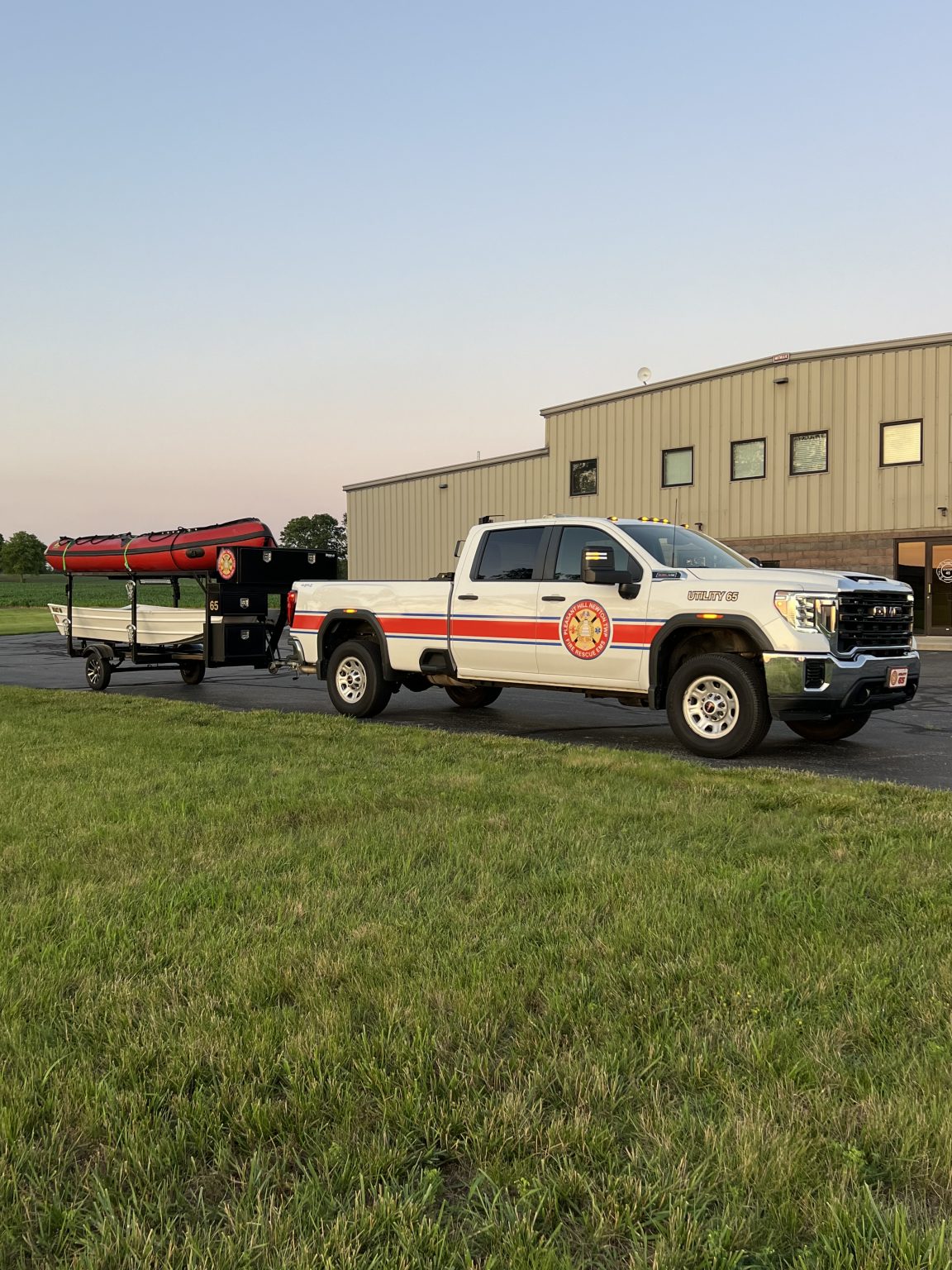 A white fire department truck with red and gold stripes is parked on pavement, towing a trailer carrying an inflatable rescue boat. A beige industrial building is in the background, and grass is in the foreground.