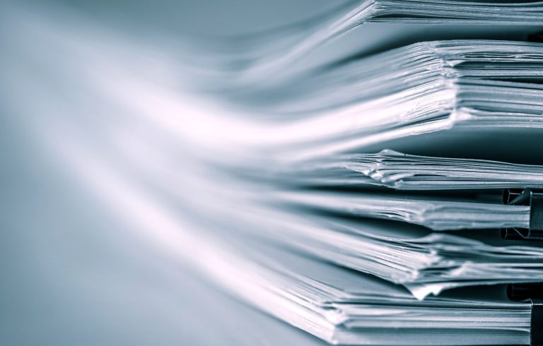 Close-up of several stacks of documents clipped together with black binder clips, papers slightly fanned out, creating a sense of organization and office work. The image has a cool blue tint.